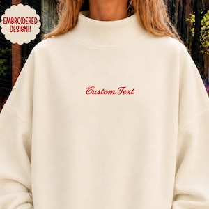 May include: An off-white, long-sleeve sweatshirt with a mock neck collar. The garment features the words Custom Text embroidered in red cursive script across the center of the chest. The design includes a scalloped badge stating Embroidered Design.