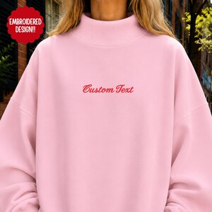 May include: A light pink mock neck sweatshirt featuring long sleeves and a relaxed fit. The center of the chest displays the red embroidered text Custom Text. A red circular badge in the upper left corner contains the text EMBROIDERED DESIGN.