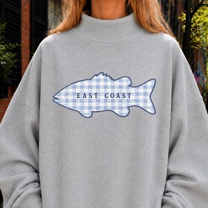 May include: A light gray mock neck sweatshirt featuring a large fish graphic on the chest. The fish is filled with a blue and white gingham pattern and contains the text EAST COAST printed in dark blue capital letters across the center.