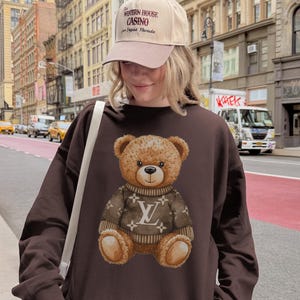 May include: A brown sweatshirt featuring a teddy bear wearing a sweater with a logo. The teddy bear is centered on the sweatshirt. The person in the image is wearing a beige baseball cap with text.