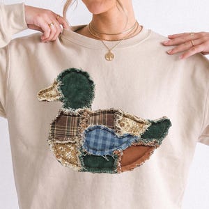 May include: A beige crewneck sweatshirt featuring a large, central duck-shaped appliqué. The duck is constructed from various fabric patches, including dark green corduroy, brown plaid, blue gingham, and floral patterns with frayed edges.