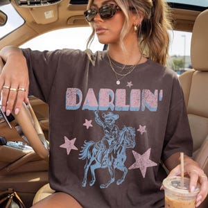 Puede incluir: Camiseta marr&oacute;n con la palabra "Darlin'" en rosa y azul, un gr&aacute;fico de un vaquero a caballo y estrellas rosas. La camiseta tiene un corte holgado y casual.