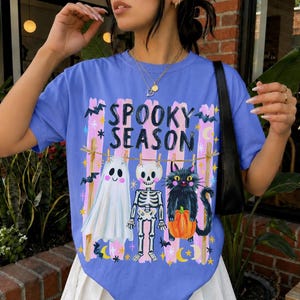 May include: A periwinkle blue t-shirt featuring a graphic print with the text "SPOOKY SEASON" above illustrations of a white ghost, a skeleton, and a black cat holding an orange pumpkin, all hanging from a clothesline with bats and stars.