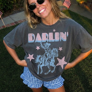 Puede incluir: Una camiseta gris carb&oacute;n con la palabra "DARLIN'" en rosa y azul, y un dibujo de un vaquero montando a caballo. La persona lleva un pantal&oacute;n corto a cuadros azul y blanco y botas rojas. Hay banderas americanas en el fondo.
