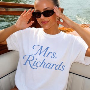 Może przedstawiać: Biały t-shirt z kr&oacute;tkim rękawem z napisem Mrs. Richards wydrukowanym jasnoniebieską kursywą na klatce piersiowej. Odzież ma okrągły dekolt i standardowy kr&oacute;j, wykonana z gładkiego białego materiału z niebieskimi literami.