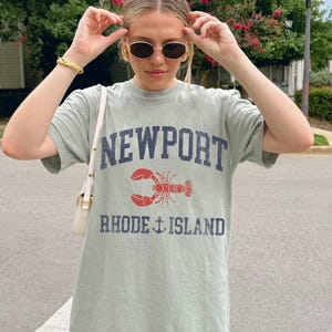 May include: Sage green t-shirt with the words "NEWPORT" and "RHODE ISLAND" printed in navy blue. A red lobster graphic is centered between the words. The shirt is a casual, oversized fit.