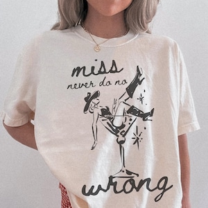 May include: Cream-colored t-shirt featuring a black graphic of a woman in a martini glass, wearing a cowboy hat and boots. The text "miss never do no wrong" is printed above and below the illustration in a handwritten style.