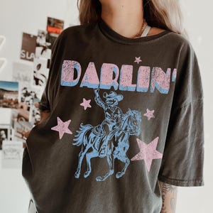 Puede incluir: Camiseta gris oscuro con la palabra "DARLIN'" en letras rosas y azules. Debajo, un gr&aacute;fico azul y rosa de un vaquero a caballo, con estrellas rosas. La camiseta tiene un aspecto vintage y desgastado, probablemente de algod&oacute;n.