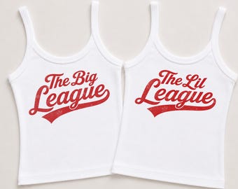 Może przedstawiać: Dwie białe koszulki bez rękawów z okrągłym dekoltem. Lewa koszulka ma czerwony napis The Big League, a prawa koszulka ma czerwony napis The Lil League. Oba nadruki charakteryzują się celowo postarzaną, przetartą teksturą.