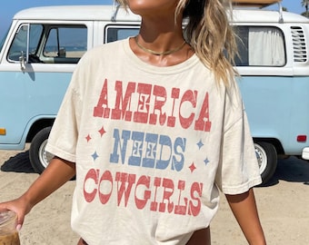 May include: Cream-colored t-shirt with the text "AMERICA NEEDS COWGIRLS" in red and blue. The shirt has a vintage, distressed look. A surfboard is on top of a light blue van in the background.