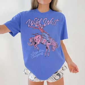 May include: A blue t-shirt featuring a pink graphic of a cowgirl riding a horse. The words "Wild West" and "Long live Cowgirls" are written in pink script on the shirt. The shirt has short sleeves.