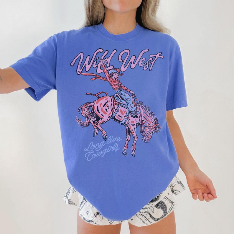 May include: A blue t-shirt featuring a pink graphic of a cowgirl riding a horse. The words "Wild West" and "Long live Cowgirls" are written in pink script on the shirt. The shirt has short sleeves.