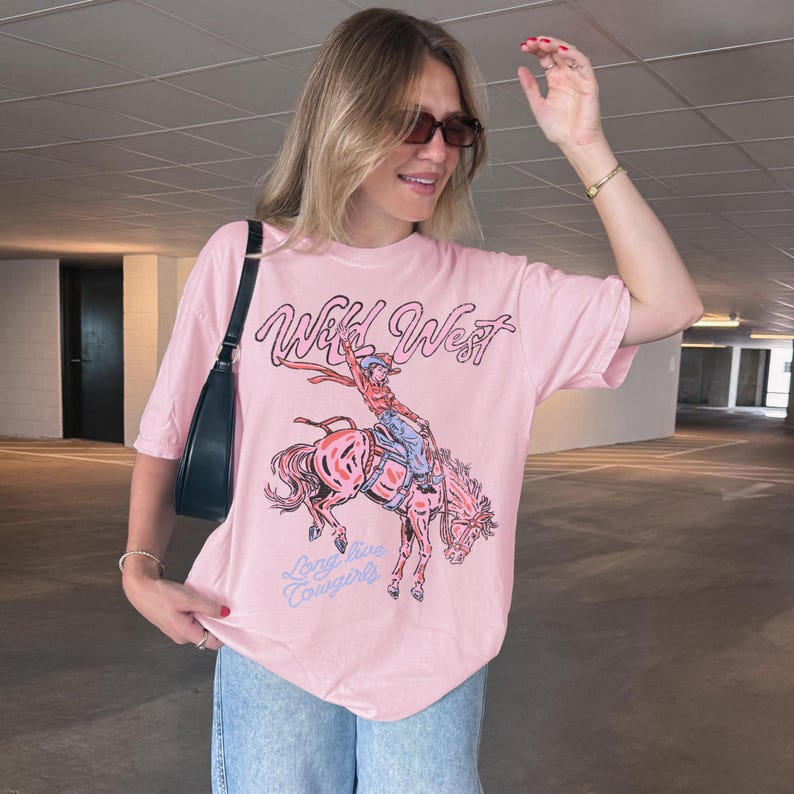 May include: A light pink t-shirt with a graphic of a cowboy riding a horse, the words "Wild West" and "Long live Cowgirls" are printed on the shirt. The shirt is made of a soft material.