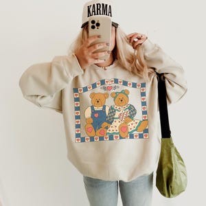 May include: Beige sweatshirt featuring a graphic of two teddy bears within a heart-bordered frame. The bears are wearing overalls and a dress. A white cap with the word "KARMA" is visible.