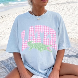 May include: Light blue t-shirt with the word "LATER" in pink gingham letters and a green alligator graphic. The shirt is a relaxed fit and suitable for casual wear.