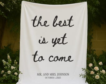 May include: A white fabric banner with the text "the best is yet to come" in black script. Below the text is "MR. AND MRS. JOHNSON OCTOBER 1, 2025". The banner is displayed outdoors with greenery and flowers in the background.