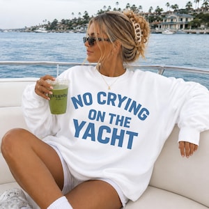 May include: White sweatshirt with the text "NO CRYING ON THE YACHT" in blue. The person is wearing sunglasses and holding a green drink. The background shows a body of water and buildings.