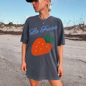 May include: Dark grey oversized t-shirt with a large orange strawberry graphic and green leaves. The blue text reads "La Fraise" above the strawberry, with "100% Organic" below. The shirt has short sleeves.