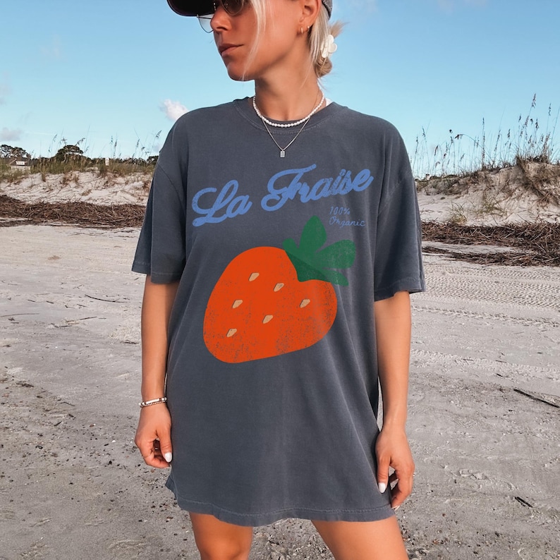 May include: Dark grey oversized t-shirt with a large orange strawberry graphic and green leaves. The blue text reads "La Fraise" above the strawberry, with "100% Organic" below. The shirt has short sleeves.