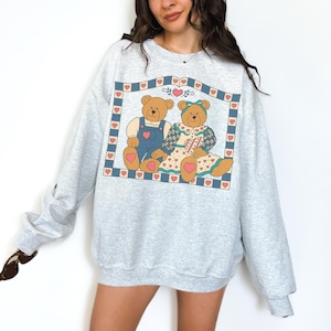 May include: A light gray sweatshirt featuring a graphic of two teddy bears within a heart-bordered frame. One bear wears blue overalls, the other a dress. The design includes hearts and a blue and pink color scheme.