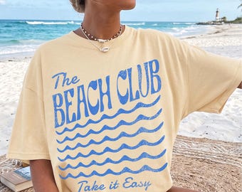 May include: A pale yellow t-shirt featuring the words "The Beach Club" in blue, above blue wave graphics. The phrase "Take it Easy" is printed below. The shirt is worn on a sandy beach.