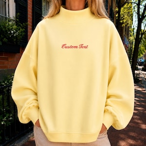 May include: A pale yellow mock neck sweatshirt featuring long sleeves with dropped shoulders and ribbed cuffs. The center of the chest displays the words Custom Text embroidered in a red cursive font, creating a simple and clean aesthetic.