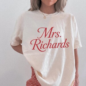 Może przedstawiać: Kremowy t-shirt z kr&oacute;tkim rękawem z czerwonym napisem "Mrs. Richards" wydrukowanym kursywą na klatce piersiowej. Koszulka ma okrągły dekolt i luźny kr&oacute;j, zestawiona z czerwono-białymi spodniami w kratę.