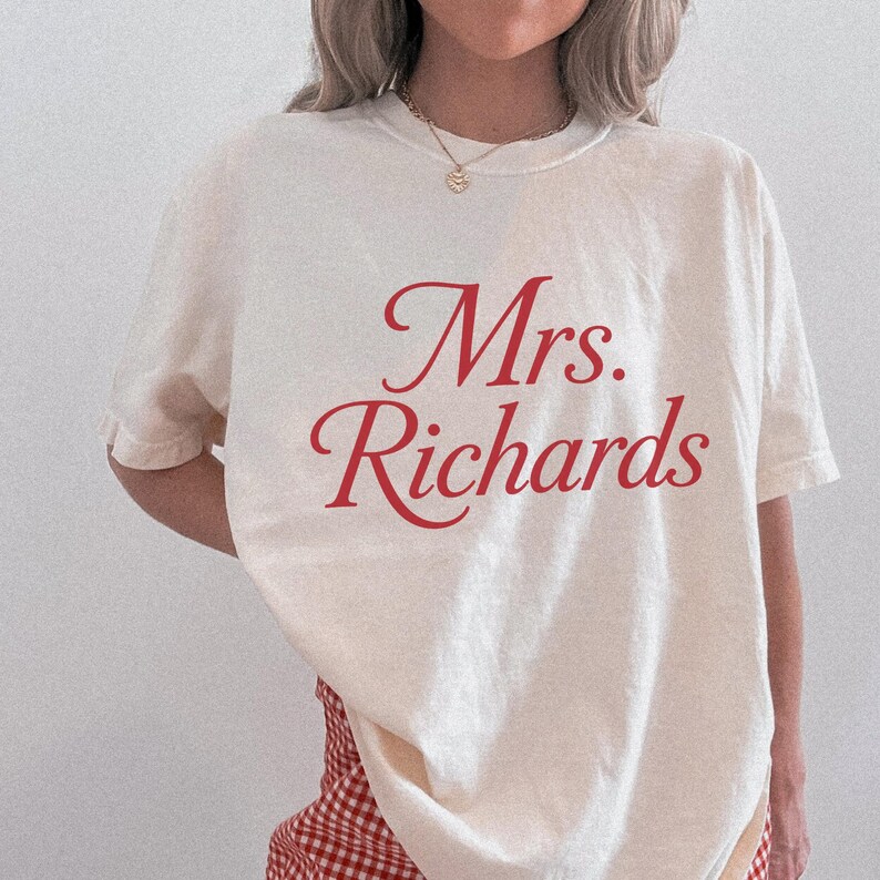 Może przedstawiać: Kremowy t-shirt z kr&oacute;tkim rękawem z czerwonym napisem "Mrs. Richards" wydrukowanym kursywą na klatce piersiowej. Koszulka ma okrągły dekolt i luźny kr&oacute;j, zestawiona z czerwono-białymi spodniami w kratę.