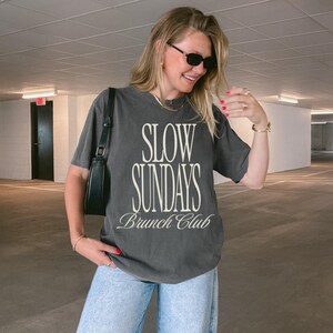 Peut inclure: T-shirt gris fonc&eacute; avec l'inscription "SLOW SUNDAYS Brunch Club" en blanc. Le t-shirt est port&eacute; avec un jean bleu clair et un sac &agrave; bandouli&egrave;re noir. La personne porte des lunettes de soleil et des bijoux.