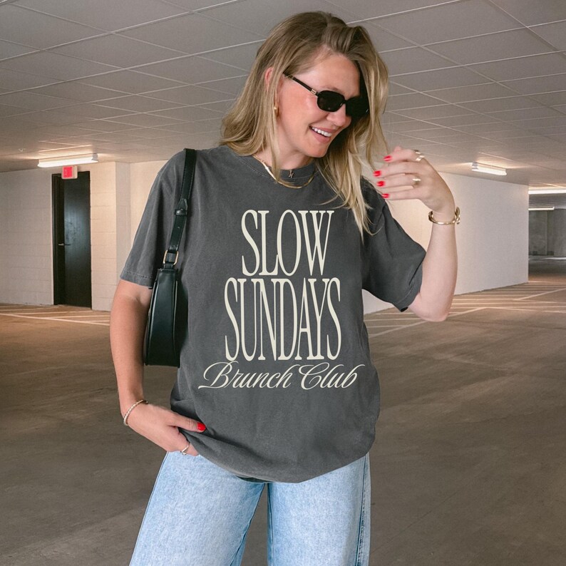 Peut inclure: T-shirt gris fonc&eacute; avec l'inscription "SLOW SUNDAYS Brunch Club" en blanc. Le t-shirt est port&eacute; avec un jean bleu clair et un sac &agrave; bandouli&egrave;re noir. La personne porte des lunettes de soleil et des bijoux.