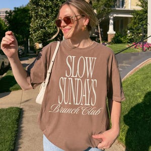Peut inclure: Un t-shirt marron avec l'inscription blanche "SLOW SUNDAYS Brunch Club". Le t-shirt est port&eacute; avec des lunettes de soleil et un sac blanc. L'image est prise en ext&eacute;rieur par une journ&eacute;e ensoleill&eacute;e.