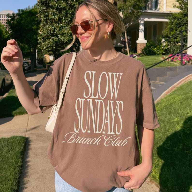 Peut inclure: Un t-shirt marron avec l'inscription blanche "SLOW SUNDAYS Brunch Club". Le t-shirt est port&eacute; avec des lunettes de soleil et un sac blanc. L'image est prise en ext&eacute;rieur par une journ&eacute;e ensoleill&eacute;e.