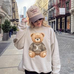 May include: Beige sweatshirt featuring a brown teddy bear wearing a sweater with a white logo. The person is wearing a beige and brown baseball cap with the text "The Western House Casino Las Vegas Nevada".