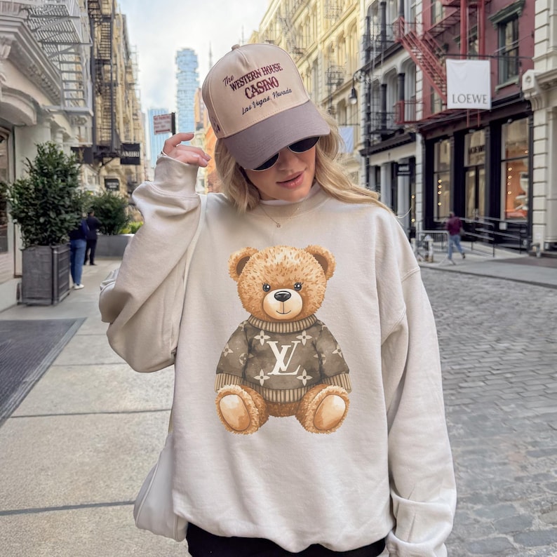 May include: Beige sweatshirt featuring a brown teddy bear wearing a sweater with a white logo. The person is wearing a beige and brown baseball cap with the text "The Western House Casino Las Vegas Nevada".