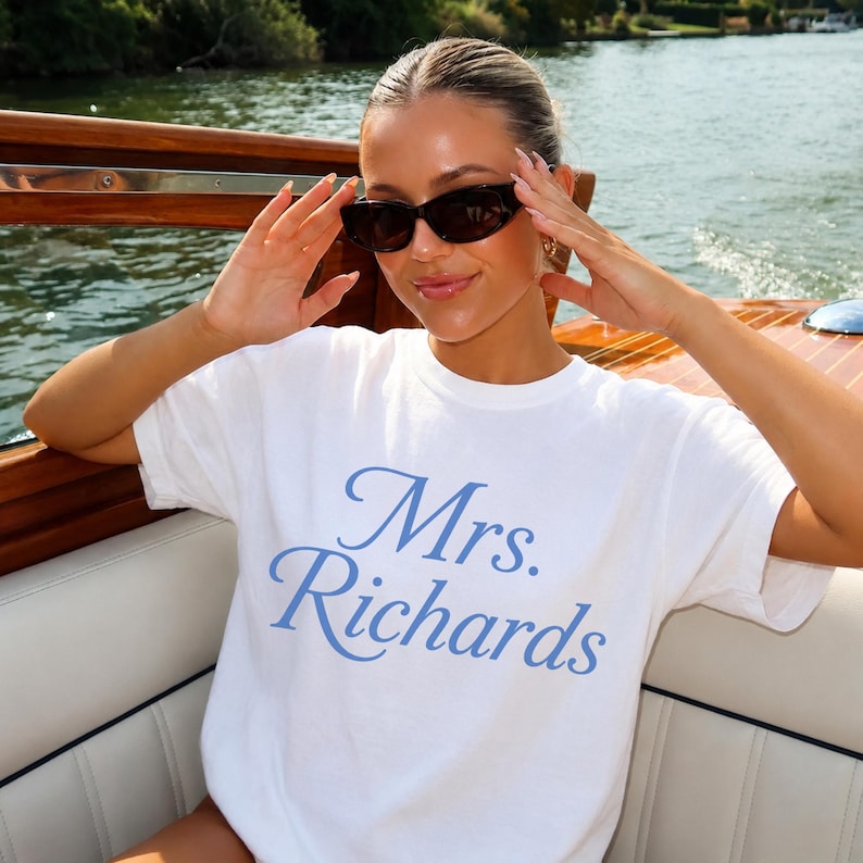 Może przedstawiać: Biały t-shirt z kr&oacute;tkim rękawem z napisem Mrs. Richards wydrukowanym jasnoniebieską kursywą na klatce piersiowej. Odzież ma okrągły dekolt i standardowy kr&oacute;j, wykonana z gładkiego białego materiału z niebieskimi literami.