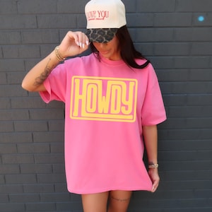 May include: A vibrant pink t-shirt featuring the word "HOWDY" in yellow block letters. The person is wearing a cream baseball cap with the words "LOVE YOU" in red and a leopard print brim.