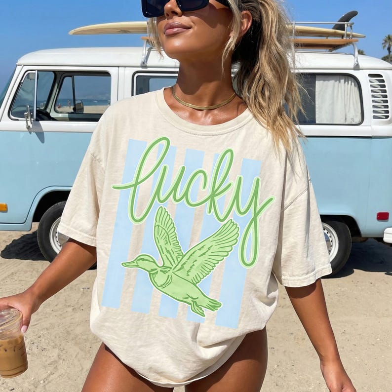 May include: Cream-colored t-shirt with the word "Lucky" in green script and a green duck graphic. The design is set against a light blue striped background. The shirt is a casual, short-sleeved style.