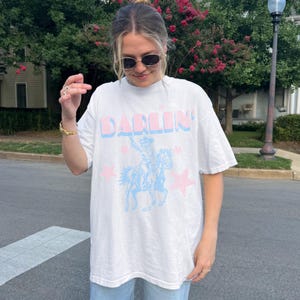 Puede incluir: Camiseta blanca con la palabra "DARLIN'" en letras rosas y azules. Un vaquero azul a caballo se muestra con estrellas rosas. La persona lleva gafas de sol.