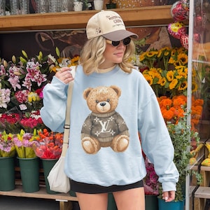 May include: A light blue sweatshirt featuring a brown teddy bear wearing a brown sweater with a white logo. The person is wearing a tan baseball cap with dark sunglasses. The background includes a flower shop with various colorful flowers.