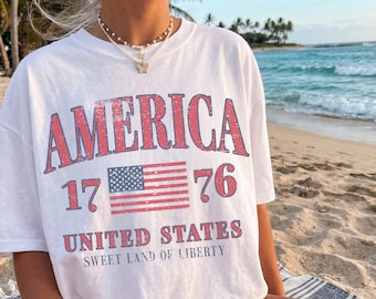 Può includere: T-shirt bianca con la scritta "AMERICA" in rosso, il numero "1776" e una bandiera americana. Sotto la bandiera, il testo recita "UNITED STATES SWEET LAND OF LIBERTY". La maglietta è indossata su una spiaggia.