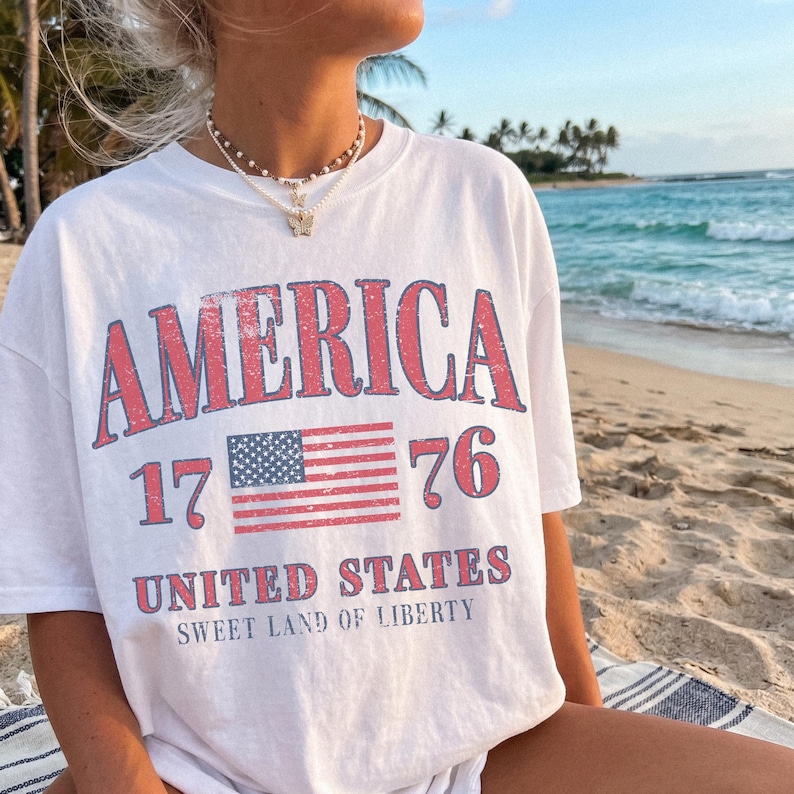 May include: White t-shirt featuring the word "AMERICA" in red, the number "1776", and a US flag graphic. Below the flag, the text reads "UNITED STATES SWEET LAND OF LIBERTY". The shirt is worn on a beach.