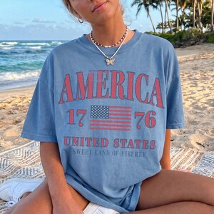 May include: A blue t-shirt featuring the word "AMERICA" in red, with the number "1776" and an American flag graphic. The text "UNITED STATES" and "SWEET LAND OF LIBERTY" are printed below. The shirt is a casual style.