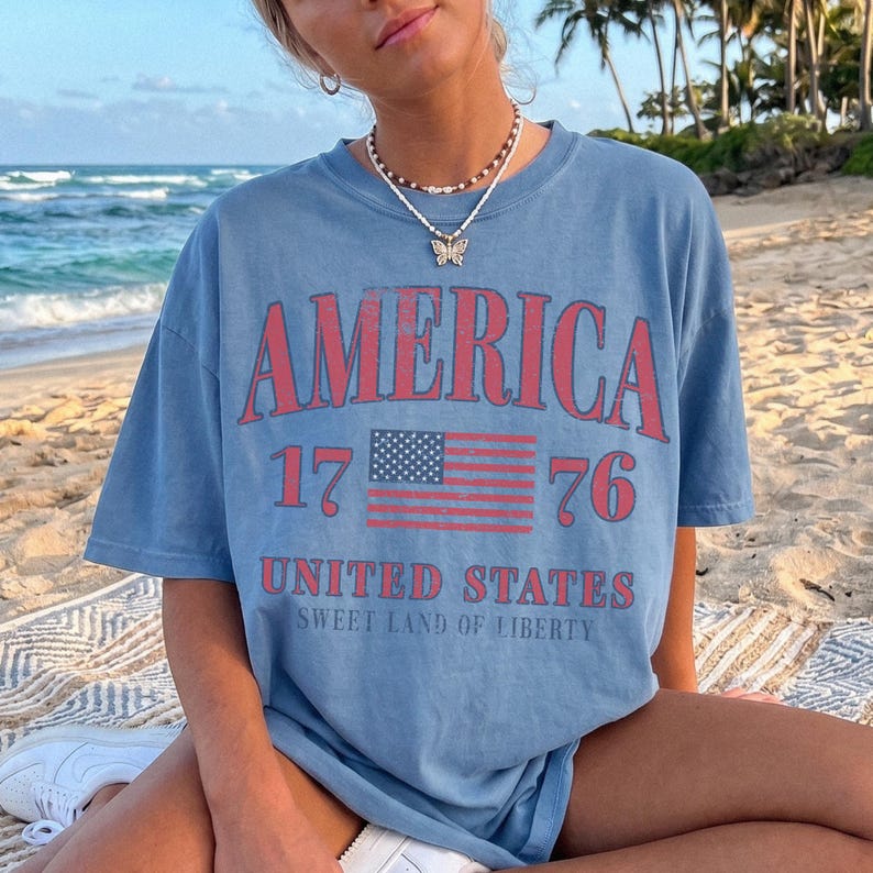 May include: A blue t-shirt featuring the word "AMERICA" in red, with the number "1776" and an American flag graphic. The text "UNITED STATES" and "SWEET LAND OF LIBERTY" are printed below. The shirt is a casual style.
