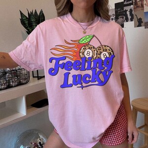 K&ouml;nnte beinhalten: Hellrosa T-Shirt mit dem Schriftzug "Feeling Lucky" in blauer Schrift. Die Grafik zeigt zwei Kirschen mit Leopardenmuster und der Zahl 8, Flammen und ein gr&uuml;nes Blatt. Ein l&auml;ssiges Kleidungsst&uuml;ck f&uuml;r den Alltag.