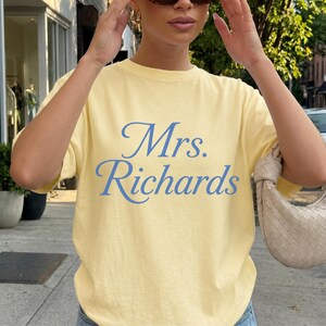 Może przedstawiać: Jasnoż&oacute;łty t-shirt z kr&oacute;tkim rękawem z napisem Mrs. Richards wydrukowanym niebieską kursywą na klatce piersiowej. Koszulka ma okrągły dekolt i luźny kr&oacute;j, co zapewnia prosty i codzienny wygląd dla każdej osoby noszącej ten ubi&oacute;r.