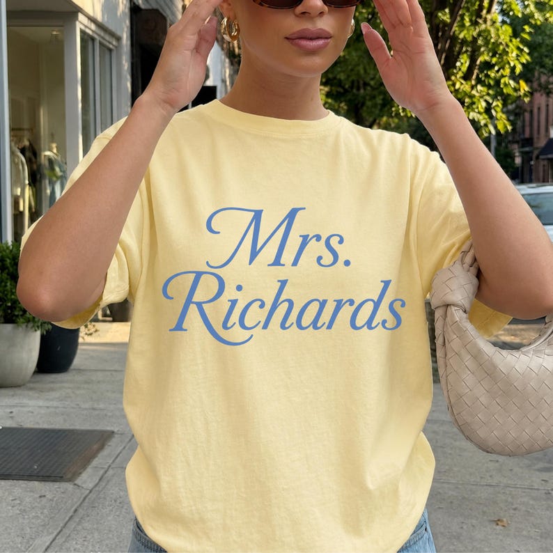 Może przedstawiać: Jasnoż&oacute;łty t-shirt z kr&oacute;tkim rękawem z napisem Mrs. Richards wydrukowanym niebieską kursywą na klatce piersiowej. Koszulka ma okrągły dekolt i luźny kr&oacute;j, co zapewnia prosty i codzienny wygląd dla każdej osoby noszącej ten ubi&oacute;r.
