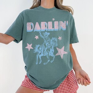 Puede incluir: Una camiseta verde azulado con la palabra "DARLIN'" en letras azules y rosas. La camiseta presenta un gr&aacute;fico de un vaquero a caballo, rodeado de estrellas rosas. La persona lleva un pantal&oacute;n corto a cuadros rojos y blancos.