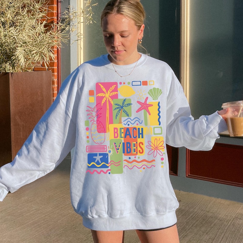 Pu&ograve; includere: Felpa bianca con una grafica colorata che dice "Beach Vibes". La grafica include immagini di palme, un ananas, una stella marina e una conchiglia. La felpa &egrave; a maniche lunghe e ha un girocollo.