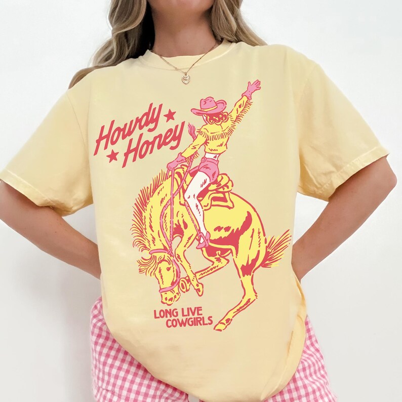 May include: Pale yellow t-shirt featuring a pink and yellow illustration of a cowgirl on a bucking bronco. The shirt displays the text "Howdy Honey" and "Long Live Cowgirls" in pink. The t-shirt has a crew neck and short sleeves.