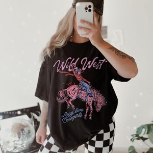 May include: Black t-shirt with a graphic of a cowgirl riding a pink horse, the words "Wild West" and "Long live Cowgirls" in pink and blue. The shirt is a relaxed fit.